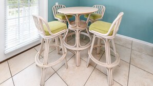 Een ronde tafel met vier stoelen, elk voorzien van een kussen. De kamer heeft een betegelde vloer en een raam met jaloezieën.