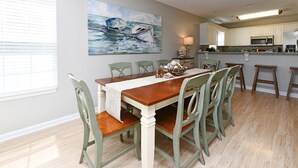A dining area with a wooden table and green chairs, a painting on the wall, and a kitchen in the background. - 2nd Row 5 BR Beach House with a Private Pool! Sale Away (Surfside Beach)