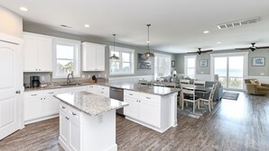 A spacious kitchen with white cabinetry, granite countertops, and a large island. There is a dining area with a table and chairs, and a view of the ocean through large windows. - Oceanfront 8 BR Beach House with a Private Pool! The Emerald (Surfside Beach)