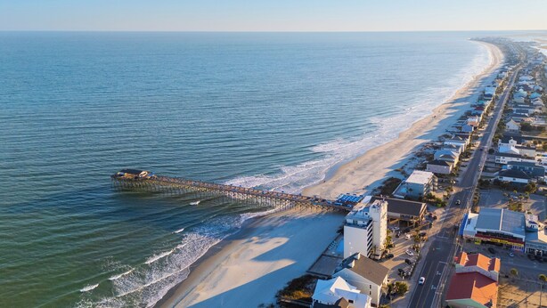 An aerial view of a coastal town with a long pier extending into the sea, adjacent to a sandy beach and lined with buildings. - Just a Walk to the Beach 5 BR Beach House with a Private Pool!  At Ease (Garden City)