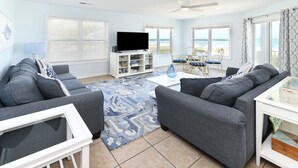 A living room with a sofa set, a coffee table, a TV, and a bookshelf. - Oceanfront 4 BR Beach House with Shared Pool! Portofino II 313B. (Surfside Beach)