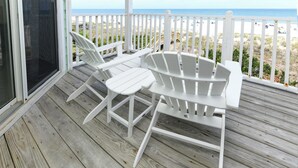 A wooden deck with white chairs and a table, overlooking a beach with a white fence and beachgoers. - Oceanfront 4 BR Beach House with Shared Pool! Portofino II 313B. (Surfside Beach)