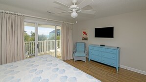 Une chambre avec un grand lit, un ventilateur de plafond, une télévision, une commode et un balcon avec des rideaux.
