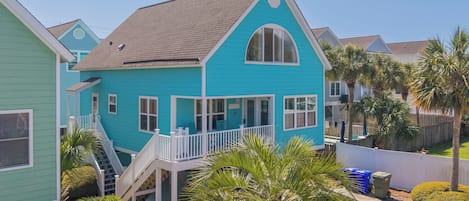 A turquoise house with a white porch, a staircase, and a fenced yard with palm trees.