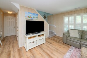 Un salon avec un téléviseur posé sur un meuble blanc, un canapé beige et un escalier orné d’un tableau.