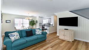 A living room with a teal sofa, wooden flooring, a TV mounted on the wall, and a kitchen in the background. - 2nd Row, 3 BR Beach House with Shared Pool in Surfside Beach! Sea Bridge 1018 (Surfside Beach)