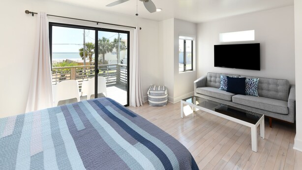 A bedroom with a large bed, a gray sofa, a flat-screen TV, and a balcony with a view of the sea. - 4 BR 2nd Row Beach House with a Private Pool! California Dreamin' (Garden City)