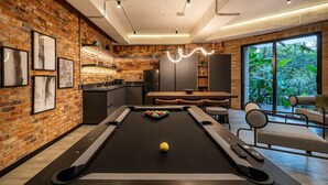 Una mesa de billar en una habitación moderna con pared de ladrillo, una zona de comedor y vistas a zonas verdes a través de amplios ventanales.