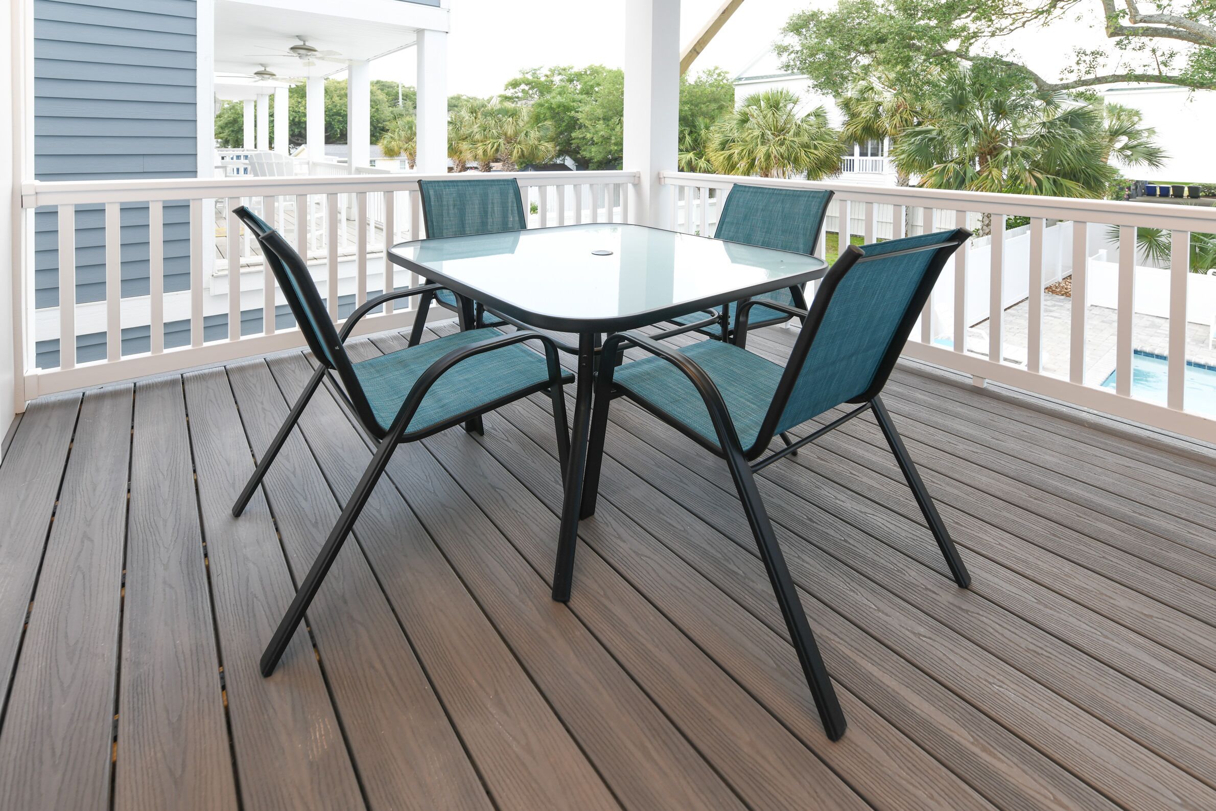 Un patio extérieur couvert, doté d’une table en verre et de quatre chaises, entouré d’une terrasse en bois et de rampes blanches.