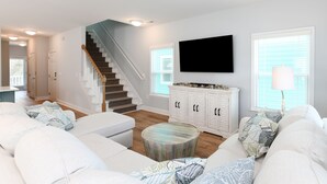 A modern living room with a staircase, white sofas, a round coffee table, and a TV mounted on the wall. - 7 BR Beach House with a Private Pool in Surfside Beach! Salty Beaches. (Surfside Beach)