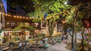 Un café en plein air avec des tables et des chaises, un arbre et un bâtiment sur lequel on peut lire l’inscription « PERGAMINO ».