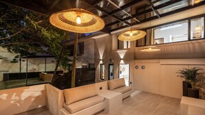 Un espace de détente moderne doté d’un haut plafond, de poutres apparentes et de grandes suspensions lumineuses en fibres tressées. On y trouve des coins salon confortables et une grande fenêtre offrant une vue sur l’extérieur.