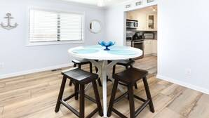 A dining area with a round table, four stools, a decorative blue vase, a wall anchor, a window with blinds, and a kitchen in the background. - 2 Bedrooms Townhouse in Surfside Beach! Dottie's Place (Surfside Beach)