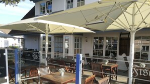 Un espace extérieur aménagé avec des tables et des chaises en bois, protégé par de grands parasols, et offrant une vue sur l’intérieur du bâtiment à travers des portes vitrées.