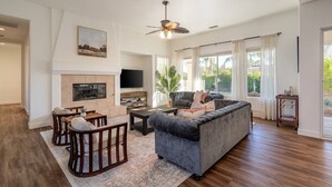 A spacious living room with a fireplace, a large sofa, a TV, and a ceiling fan. - Paradise Palms | Pool + Spa (Palm Springs)