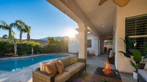 A poolside area with a wicker sofa, a table with a fire feature, and a view of a swimming pool and mountains in the background. - Paradise Palms | Pool + Spa (Palm Springs)