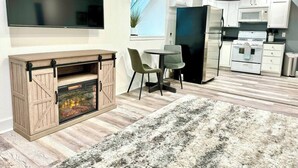 Un salon moderne doté d’une cheminée, d’un téléviseur à écran plat, d’un coin repas avec deux chaises et d’une cuisine avec des meubles et des appareils électroménagers blancs.