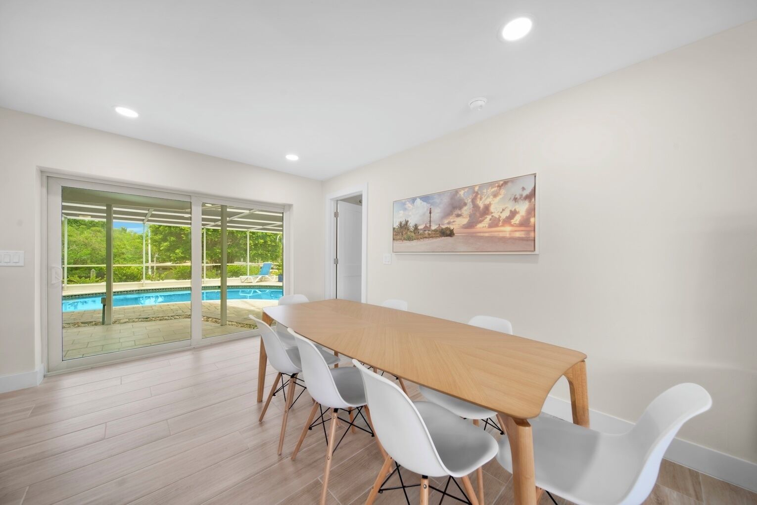 Un comedor moderno con mesa de madera y sillas blancas, ventanas amplias con vista a una piscina y follaje, y un cuadro de atardecer en la pared.