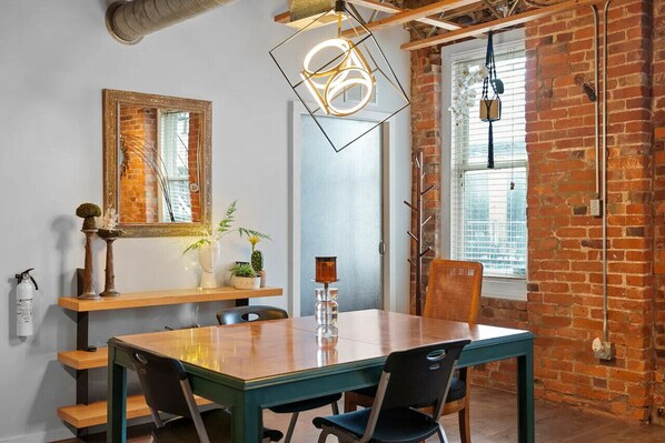 A dining area with a wooden table, chairs, a hanging light fixture, and a brick wall.
