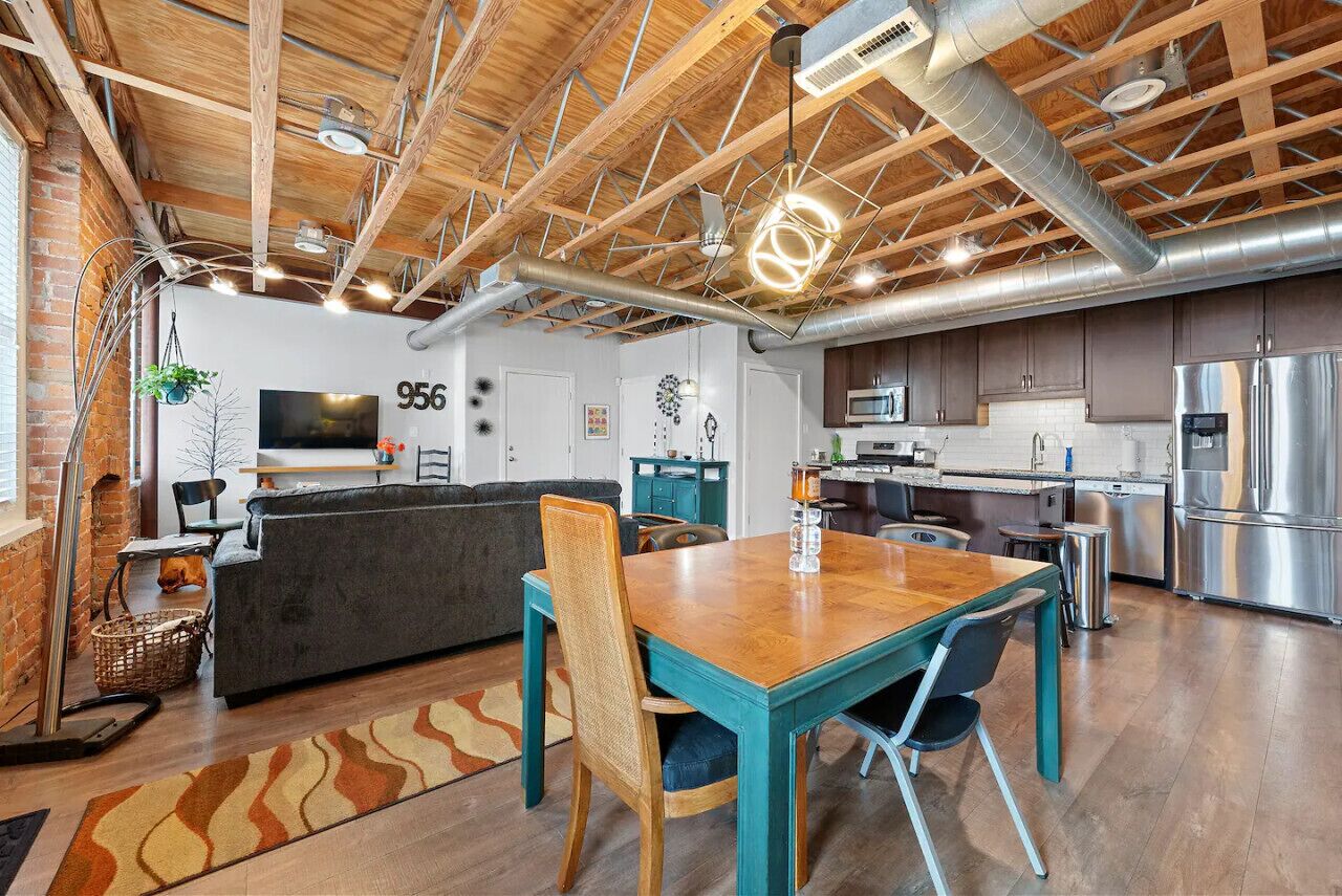 A modern kitchen with stainless steel appliances, a dining area with a wooden table and chairs, and a living area with a sofa and a TV.