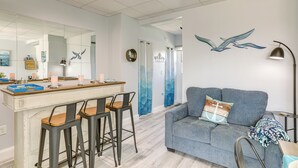 A modern living room with a grey sofa, a wooden bar stool seating area, and a large mirror reflecting the room. - Walk to Beach: Waterfront Indian Shores Condo (Indian Shores)