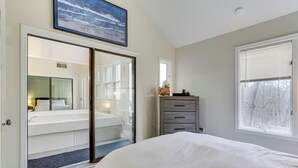 Un dormitorio con una cama grande, una puerta corrediza de vidrio que da a un baño, un aparador y una ventana con vistas a árboles.