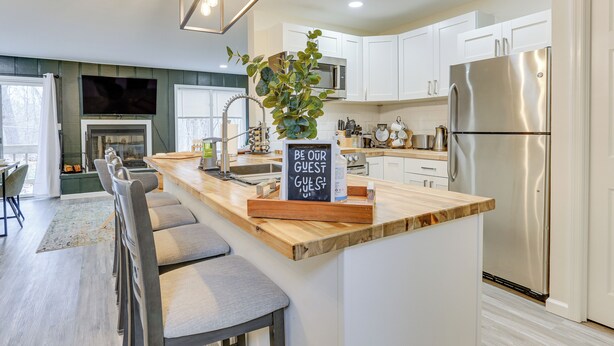 Una cocina moderna con armarios blancos, un refrigerador de acero inoxidable, una isla de madera con taburetes de bar y un letrero que dice 'BE OUR GUEST'.