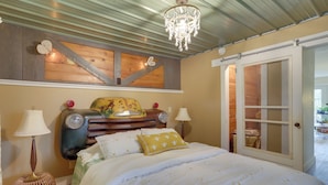 A bedroom with a vintage car headboard, a chandelier, and a sliding glass door leading to an outdoor area. - Vintage Coloma Home w/ Deck: Shop, Swim & Dine! (Coloma)