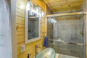 Un baño con detalles de madera, un lavabo de vidrio, un espejo con iluminación y una ducha con cerramiento de vidrio.