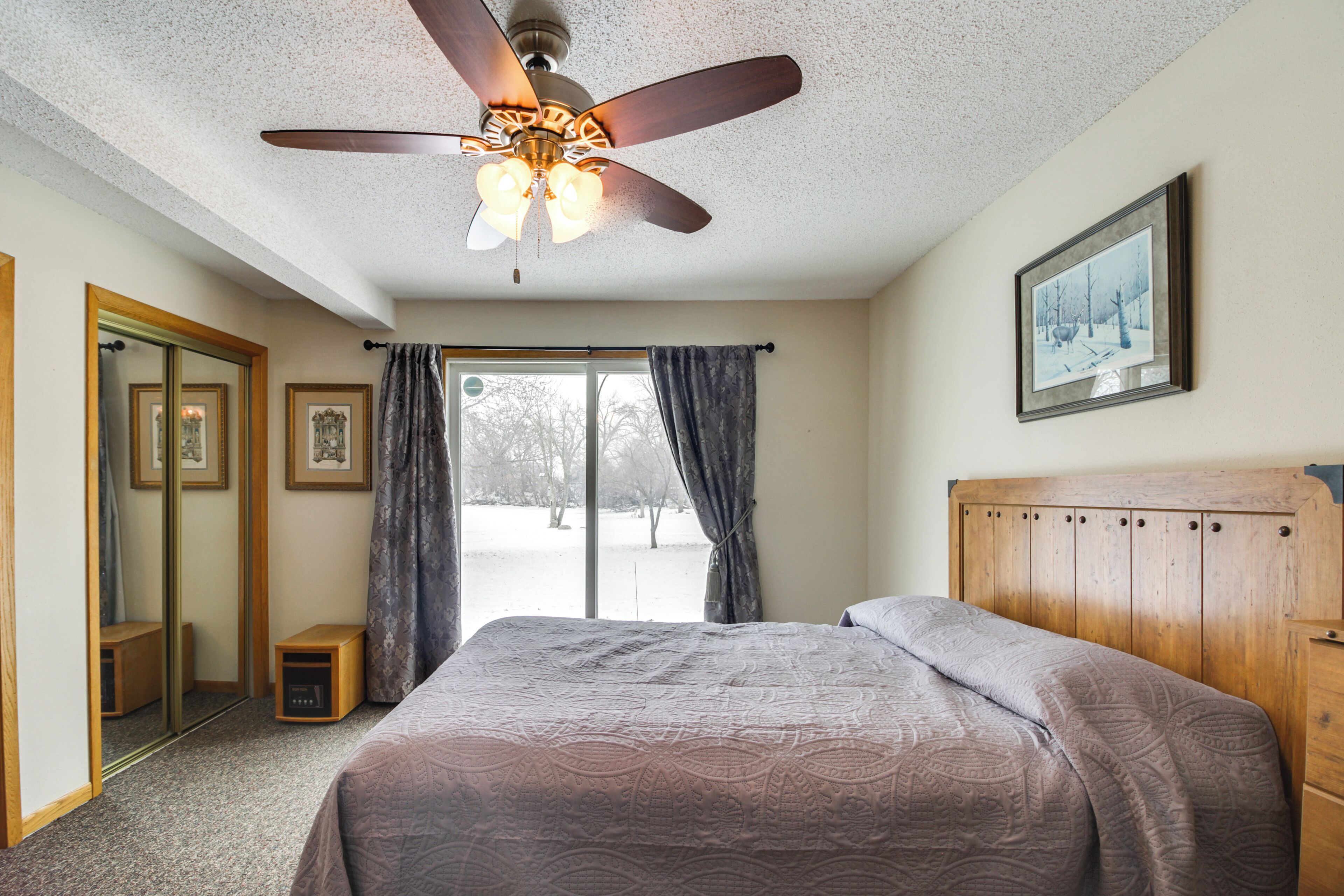 A bedroom with a bed, a ceiling fan, a window with curtains, a framed picture on the wall, and a small wooden nightstand.