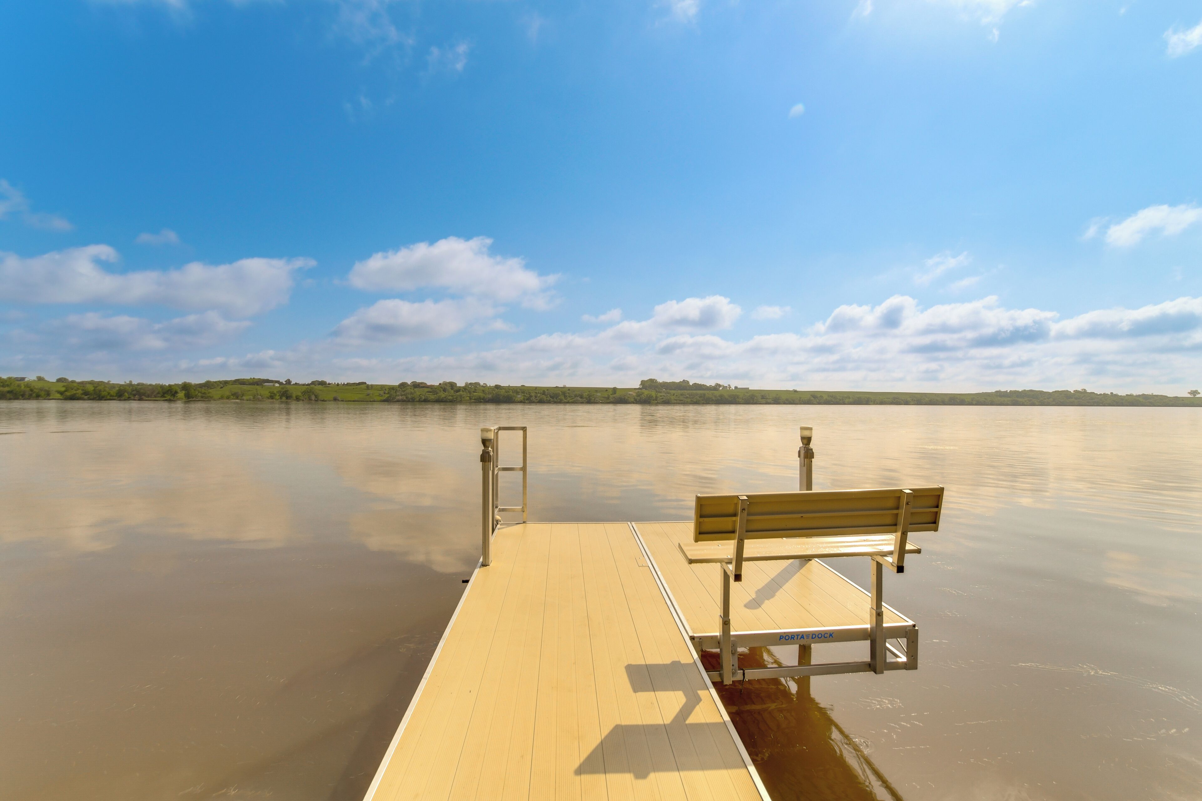 A wooden dock with a bench and a metal gate, extending over calm water with a clear sky and distant greenery.