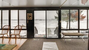 Una habitación con ventanas amplias que dan a una playa nevada, sillas de madera, un banco y un cuadro en la pared.