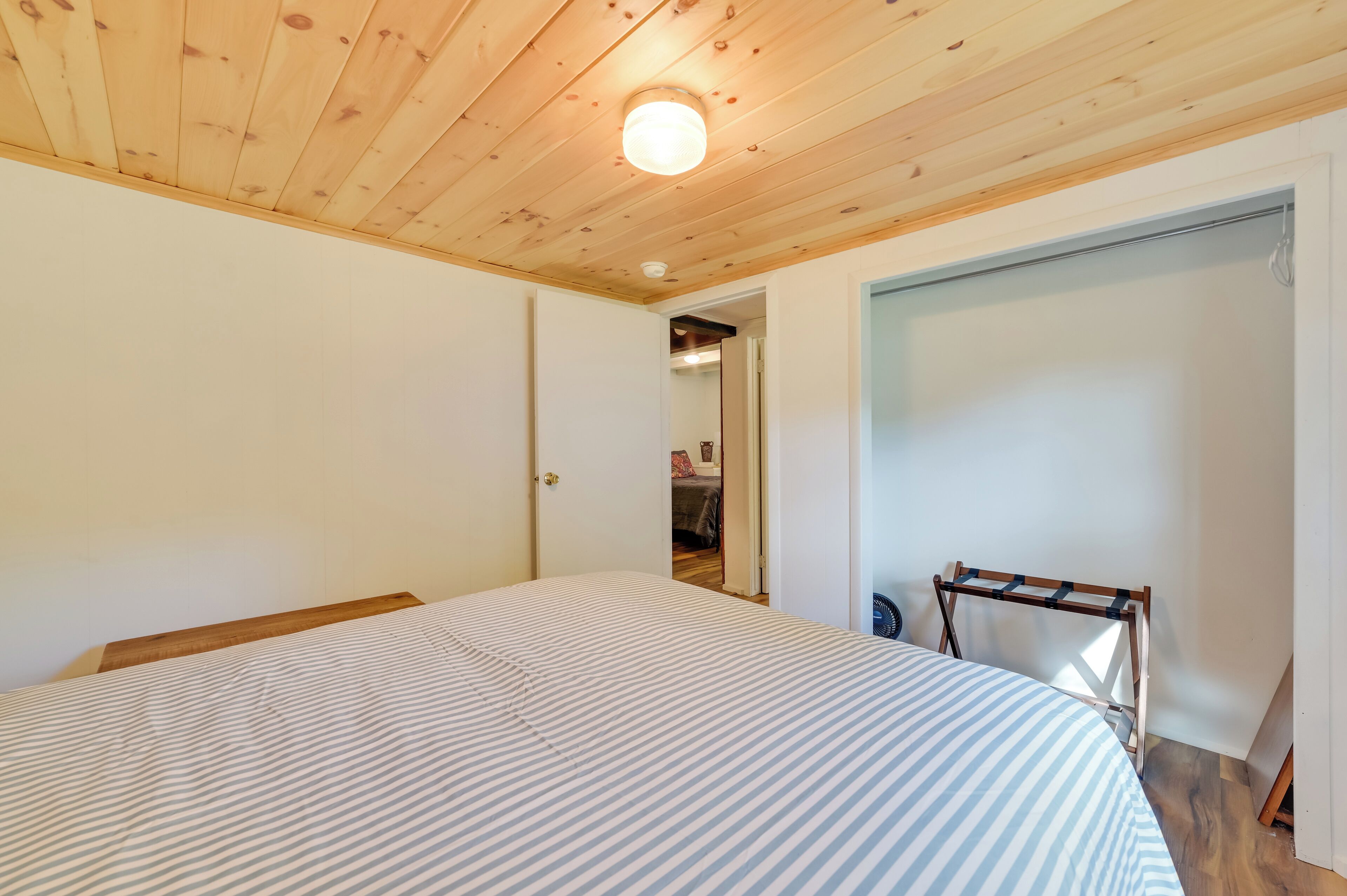 A bedroom with a wooden ceiling, a bed with striped bedding, a small table, and a door leading to another room.