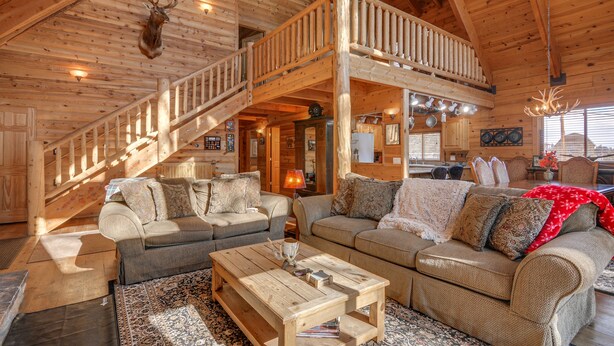 A spacious log cabin interior with a high ceiling, a staircase, and a large living area furnished with sofas and a coffee table.