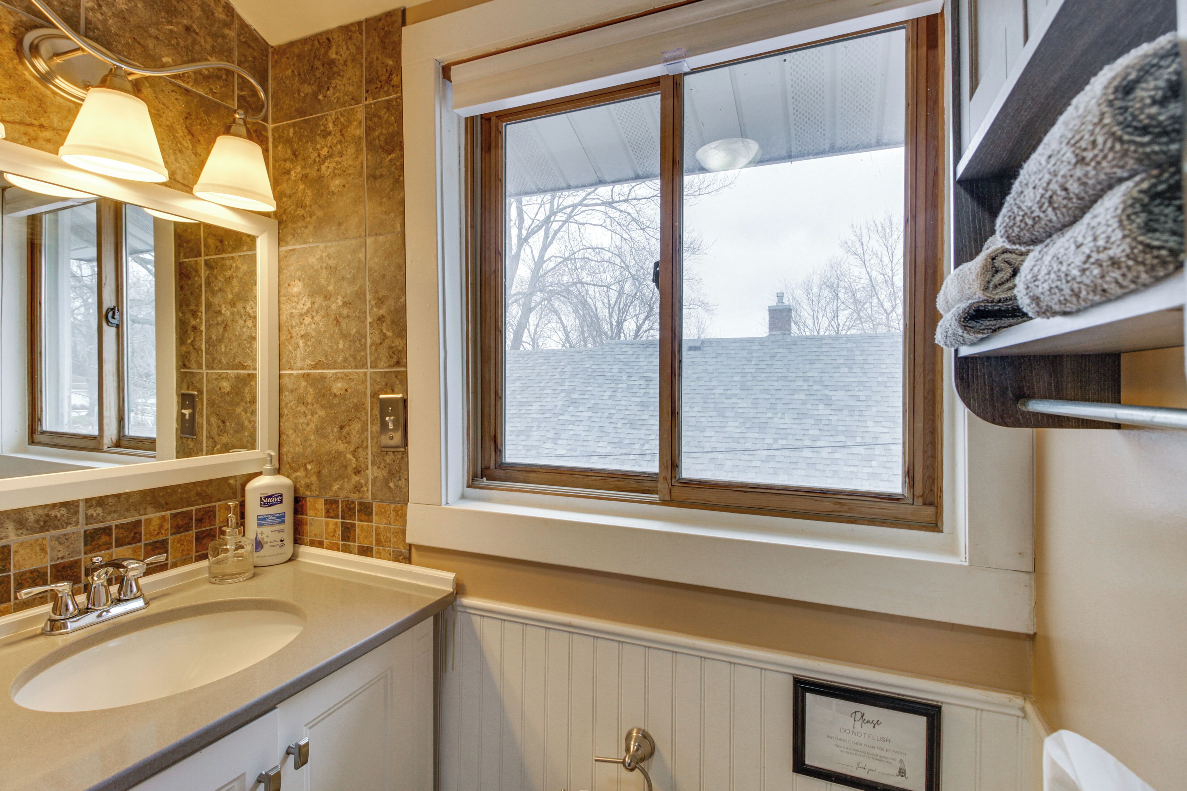 A bathroom with a large sink, mirror, and window. There is a towel rack above the sink and a framed sign on the wall.