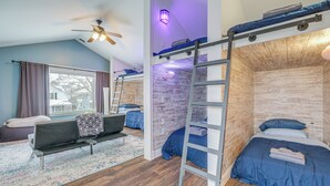 Ein schlichtes Zimmer im Schlafsaldesign mit Etagenbetten, einer Stehleiter, einem Schreibtisch und einem Fenster mit Vorhängen.