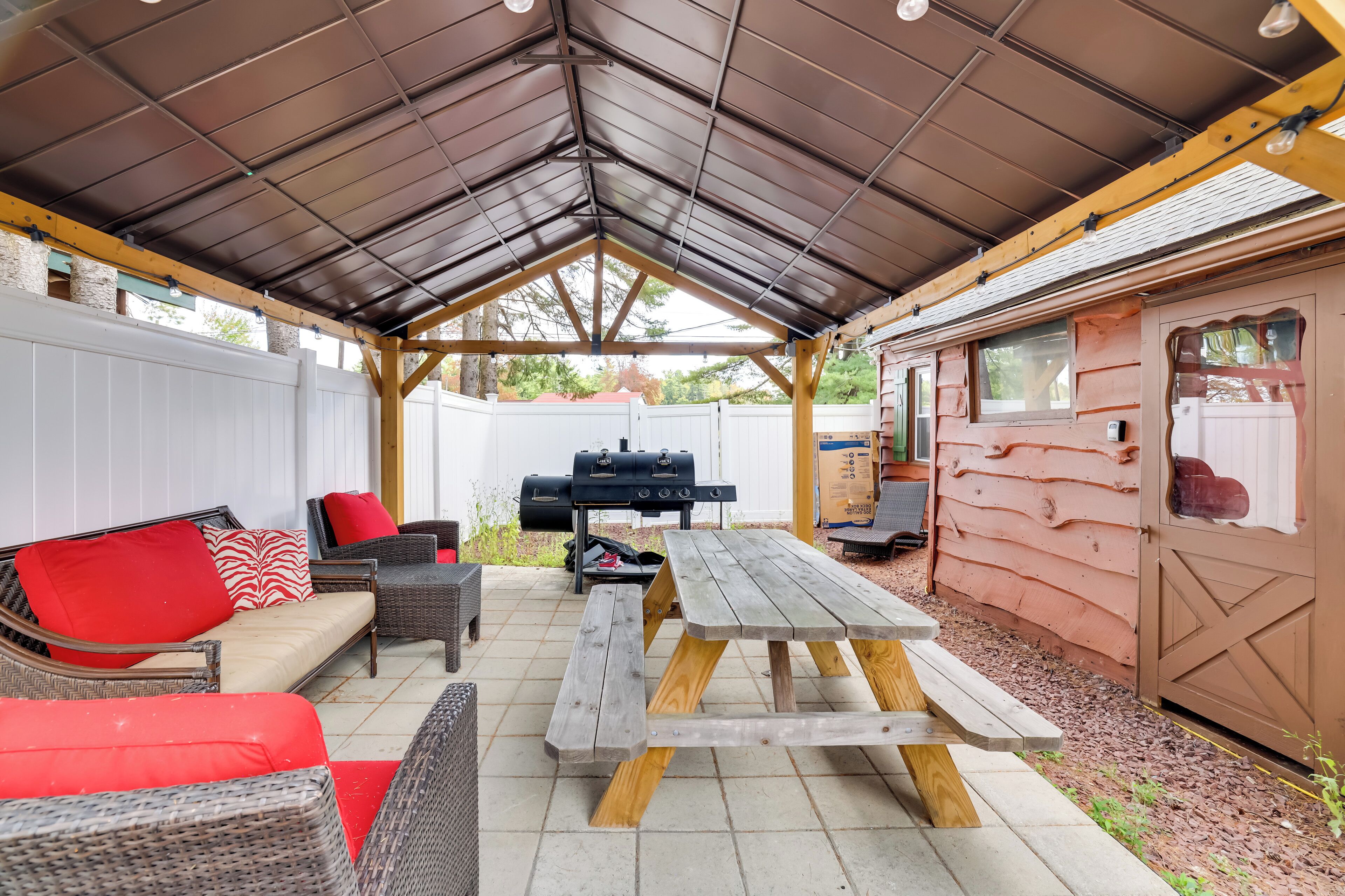 Un patio couvert avec une table et des bancs en bois, un barbecue et un bâtiment de style cabane en rondins.