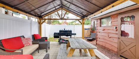 Un patio couvert avec une table et des bancs en bois, un barbecue et un bâtiment de style cabane en rondins.