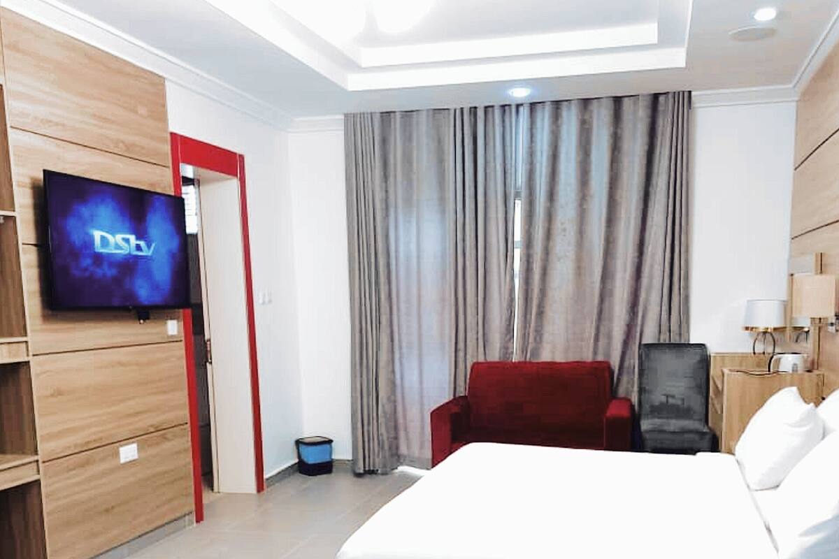 A hotel room with a bed, a TV mounted on the wall, a red chair, and a desk with a lamp.