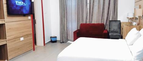 Una habitación de hotel con cama, televisión montada en la pared, una silla roja y un escritorio con lámpara.