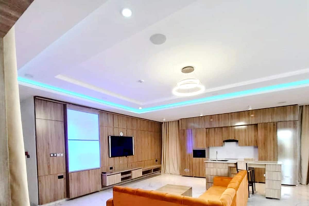 A modern living room with a large orange sofa, a flat-screen TV mounted on a wooden wall, and a kitchen area with a white countertop and cabinets.