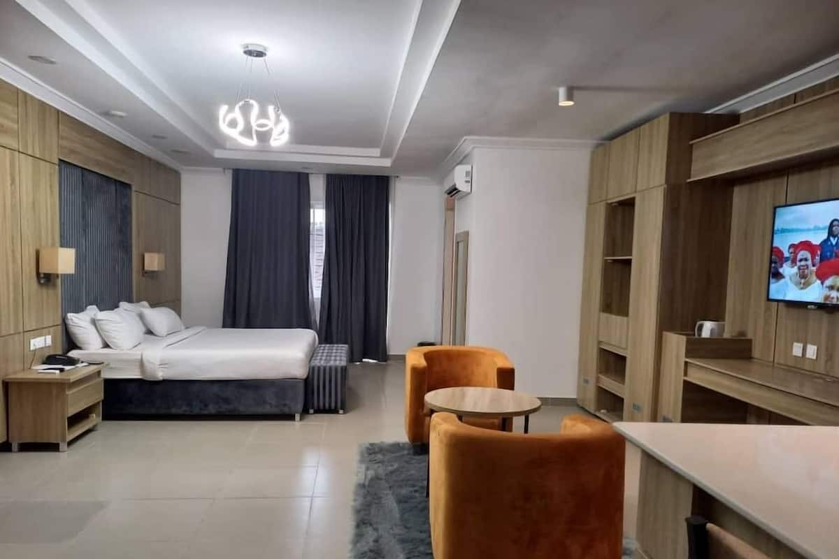 A modern hotel room with a bed, two armchairs, a small table, a TV, and wooden paneling.