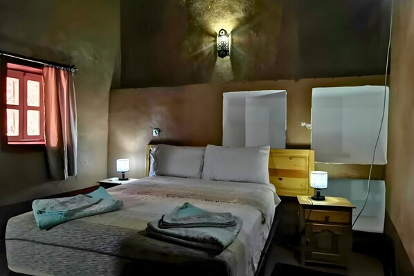 A bedroom with a bed, two bedside tables, a nightstand, a wall-mounted lamp, and a window with curtains. - Riad dar zaouia (Zagora)