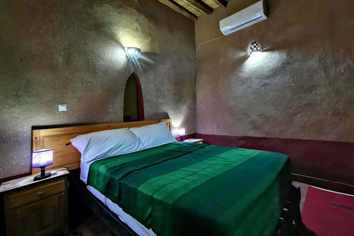 A room with a wooden headboard, a green bedspread, a nightstand with a lamp, and an air conditioning unit on the wall.