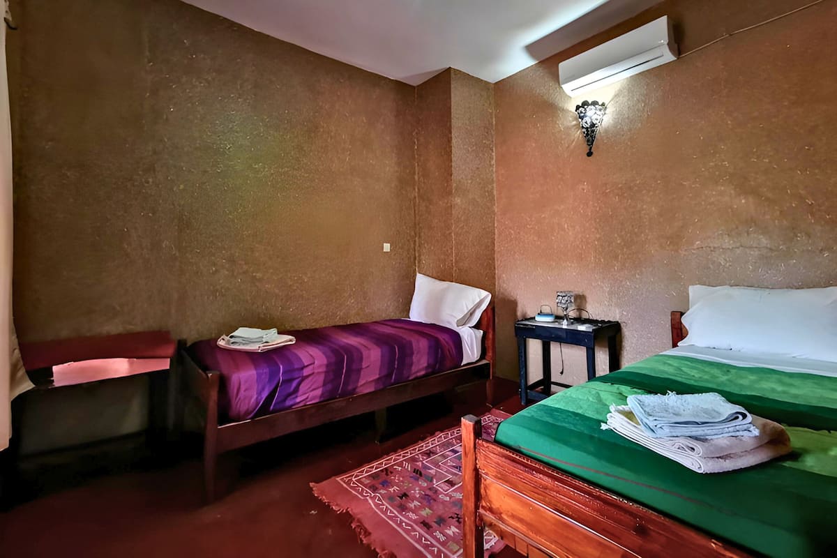 A room with two beds, one with a purple bedspread and the other with a green bedspread. There is a small table with a lamp and a wall-mounted air conditioner.