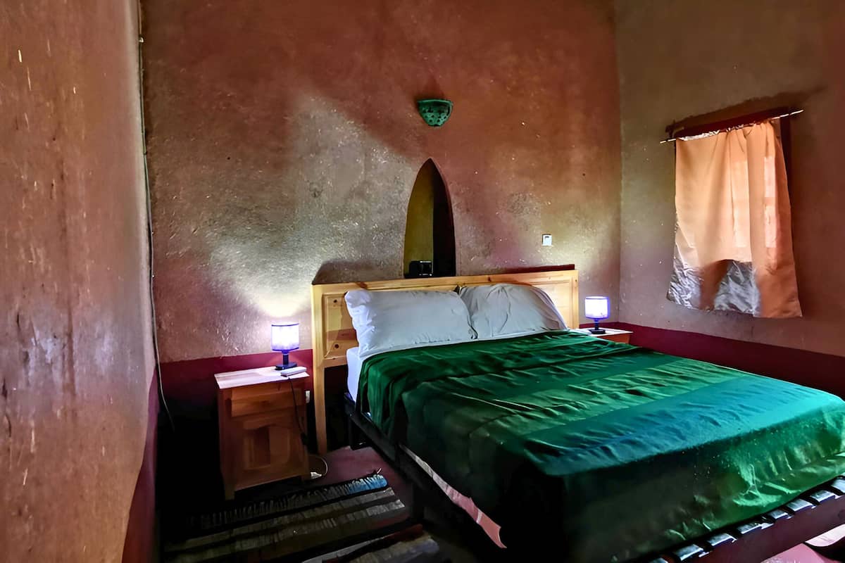 A bedroom with a bed, nightstands, a lamp, a small table, and a window with curtains.