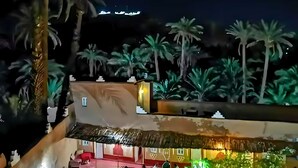 Vista nocturna de un hotel con varias edificaciones, palmeras y una estructura de techo de paja.