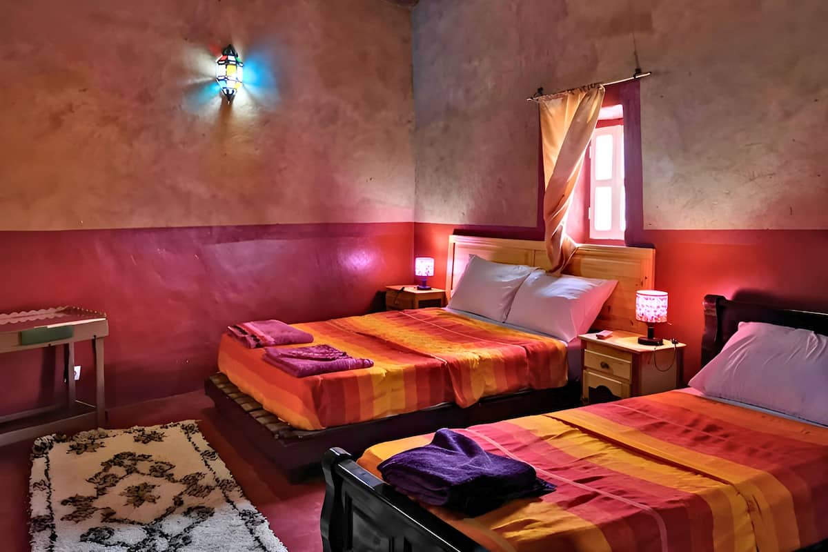 A room with two beds, each with a headboard and a nightstand. There is a small table with a lamp on one side and a window with curtains on the other.