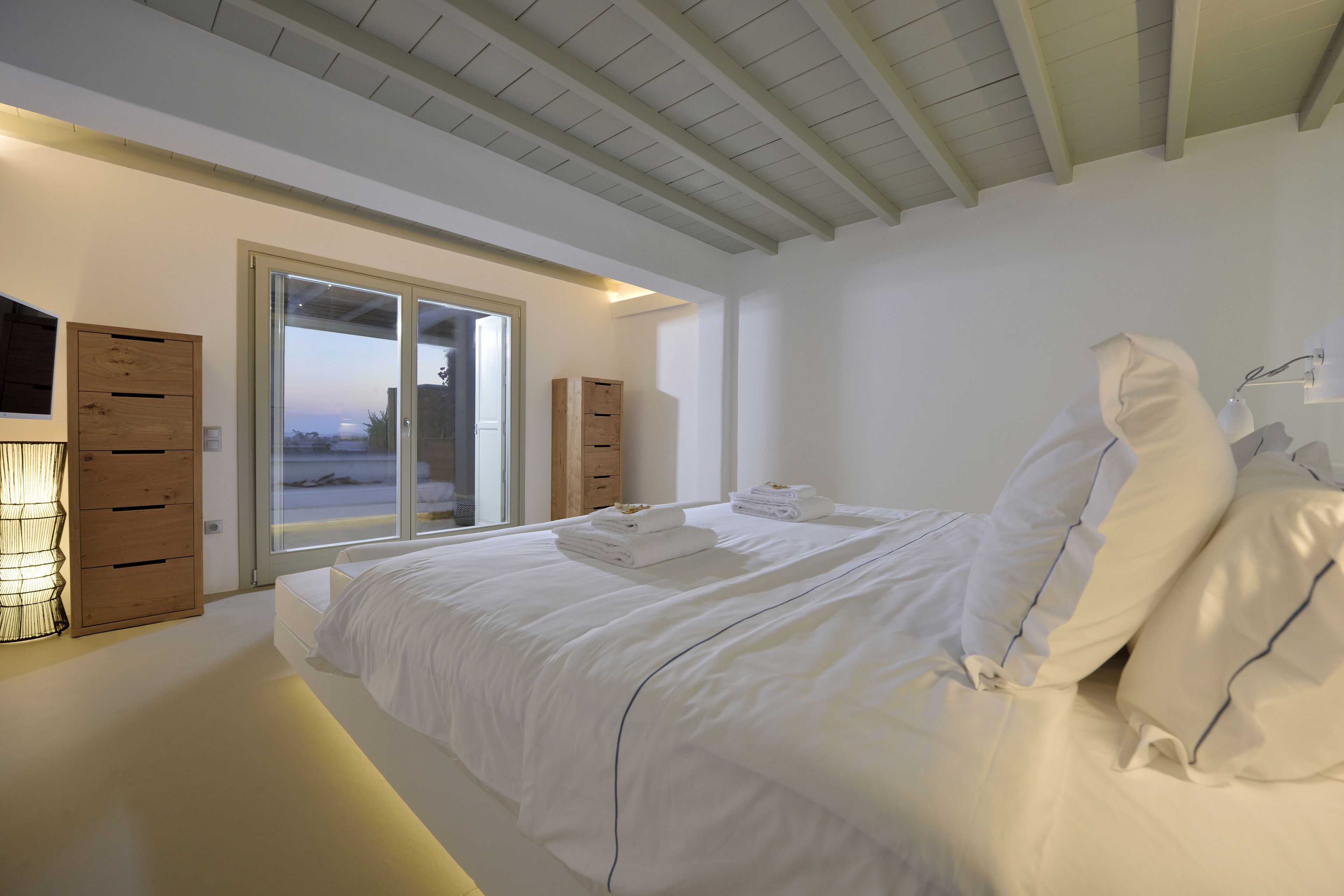 A modern bedroom with a large bed, wooden drawers, and a sliding glass door leading to a balcony.