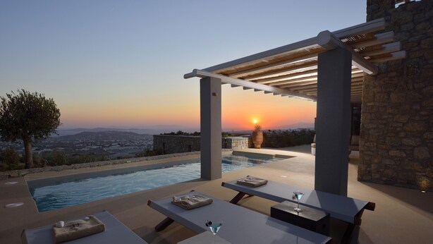 Ein Außenpoolbereich mit Liegestühlen und Tischen, einer Pergola und Blick auf den Sonnenuntergang über einer Stadtlandschaft.